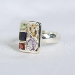 Può includere: Un anello in argento con una base rettangolare incastonata con cinque pietre preziose multicolori. Le pietre includono una piccola gemma rotonda verde, una gialla a forma di pera, una blu scura quadrata, una rossa quadrata e una ovale viola chiaro.
