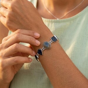 May include: A silver bracelet with a series of gemstones. The bracelet has dark blue and iridescent gray-green stones in oval and square shapes. The bracelet is worn on an arm, with a light green top visible in the background.