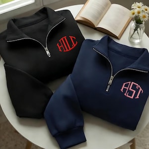 Könnte beinhalten: Zwei Pullover mit Viertelreißverschluss, einer in Schwarz und einer in Marineblau, jeweils mit Monogramm. Der schwarze Pullover hat ein rotes Monogramm, der marineblaue Pullover ein rosa Monogramm. Beide haben silberne Reißverschlüsse.