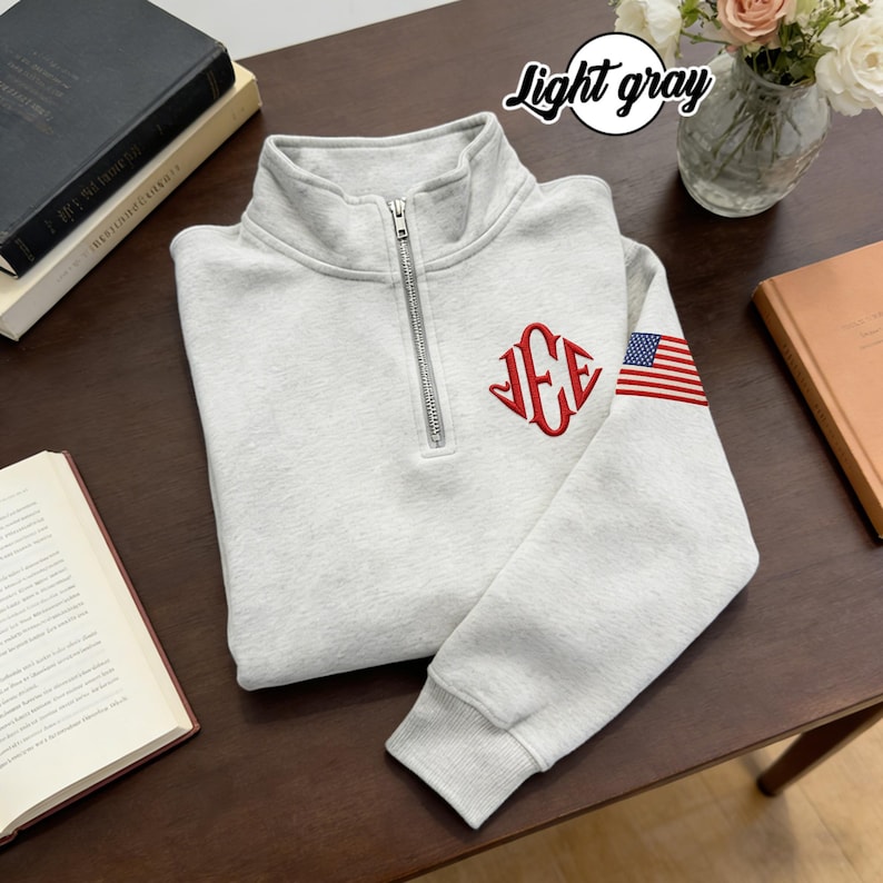 May include: Light gray quarter-zip sweatshirt with a silver zipper. The sweatshirt features a red monogram on the chest and an American flag design on the sleeve. The text "Light gray" is in a circle at the top.