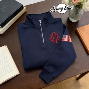 May include: A navy blue quarter-zip sweatshirt featuring a red monogram and an American flag patch on the sleeve. The sweatshirt has a silver zipper. The text "Navy blue" is in a white circle. The material appears soft.