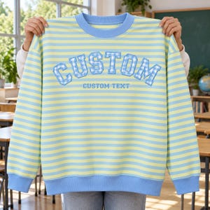 Op de afbeelding: Een sweatshirt met lange mouwen en ronde hals met horizontale lichtblauwe en lichtgele strepen. De voorkant toont het woord CUSTOM in een blauw-wit geblokt varsity-lettertype, met de tekst CUSTOM TEXT eronder gedrukt in het blauw.