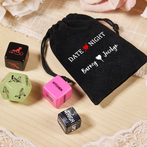 May include: A black drawstring bag with "DATE NIGHT" and names printed on it, next to several dice. The dice are black, pink, and green, with various illustrations and words like "KITCHEN" and "THIGH".