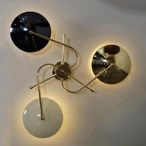 Peut inclure: Applique murale moderne avec trois luminaires circulaires. Le luminaire présente une monture en laiton et trois abat-jours circulaires : noir, doré et blanc. Chaque abat-jour est bordé de petites lumières, créant une lueur chaleureuse.