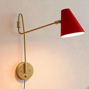 May include: A wall-mounted lamp with a red conical shade and a brass-colored adjustable arm. The lamp is attached to a round brass base with a power cord. The design is modern and minimalist.