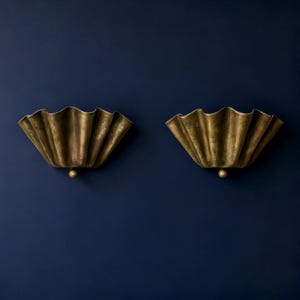 May include: Two antique brass wall sconces with a scalloped, fan-like design. Each sconce has a small, round detail at the bottom. The sconces are mounted on a dark blue wall, creating a striking contrast.