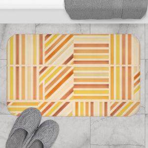 May include: A rectangular bath mat featuring a geometric pattern of vertical, horizontal, and diagonal stripes in shades of yellow, orange, and cream. The design is divided into distinct sections with varying stripe orientations and colour intensities.