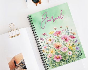 May include: A spiral-bound journal with a floral design. The cover features a watercolor illustration of pink, yellow, and purple flowers against a green background. The word "Journal" is written in pink script. Sunglasses and a gold paper clip are also visible.