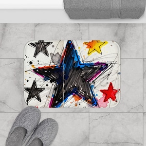 May include: A rectangular bath mat with a white background and a colorful star design. The large central star is black and blue, surrounded by smaller stars in red, yellow, and black. Splatters of color and scribbled lines add to the artistic look.