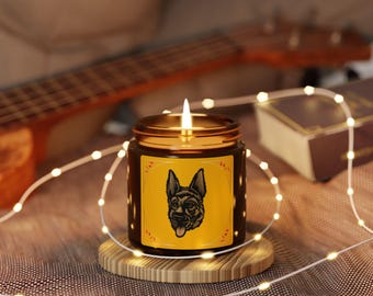 Op de afbeelding: Een brandende kaars in een bruine glazen pot met een gouden deksel. De pot heeft een geel etiket met een zwart-witte illustratie van een Duitse herdershond. De kaars staat op een houten onderzetter, met lichtslingers en een gitaar op de achtergrond.