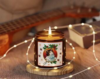 May include: A lit candle in a brown glass jar with a gold lid. The jar has a label with a cat illustration surrounded by holly and stars. The candle sits on a wooden coaster, with string lights and a ukulele in the background.