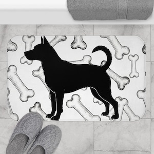 May include: A rectangular bath mat featuring a black silhouette of a dog on a white background. The background is patterned with sketched dog bones. Gray slippers are visible at the bottom left.