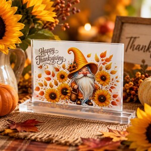 Könnte beinhalten: Ein rechteckiger, klarer Acrylblock mit einer gedruckten Illustration eines Gnoms mit gelbem Hut, umgeben von Sonnenblumen und Herbstblättern, wobei der Text Happy Thanksgiving in schwarzer Schreibschrift auf der Vorderseite steht.