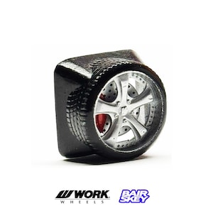 May include: A black, rectangular object with a miniature car wheel attached. The wheel has a silver rim, a red brake caliper, and a black tire. The logos "WORK WHEELS" and "BAIR SKEY" are at the bottom.