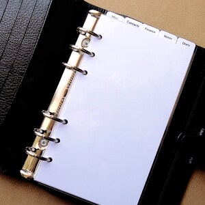May include: A black leather personal organizer with a textured finish, featuring a six-ring gold-tone metal binder mechanism. The interior contains white pages with tabbed dividers labeled Misc, Contacts, Finance, Notes, and Diary.