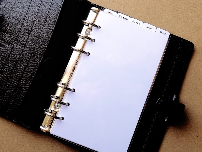 May include: A black leather personal organizer with a textured finish, featuring a six-ring gold-tone metal binder mechanism. The interior contains white pages with tabbed dividers labeled Misc, Contacts, Finance, Notes, and Diary.