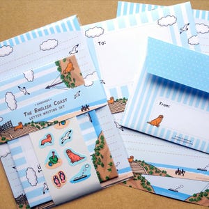 May include: A letter writing set with a coastal theme. The set includes stationery, envelopes, and stickers. The stationery features a blue and white striped design with illustrations of seagulls, clouds, and a dog. The text "The English Coast" is visible.