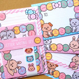 May include: A letter writing set with pink and blue stationery. The set includes envelopes, notepaper, and stickers. The designs feature cartoon animals, candy hearts, and the text "Handmade with Love" and "Letter Writing Set".