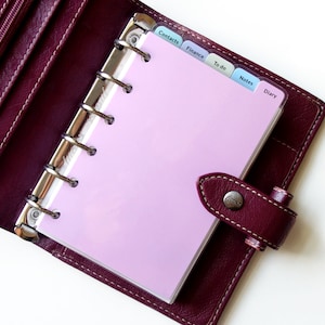 May include: A deep burgundy leather personal organizer with a six-ring metal binder mechanism. The interior contains light pink pages and translucent tabbed dividers labeled Contacts, Finance, To do, Notes, and Diary. A snap closure secures the cover.
