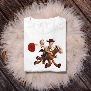 May include: White t-shirt featuring a colorful graphic of Woody and Jessie from Toy Story riding a horse. The design includes a red lasso and the characters in their signature outfits. The shirt is displayed on a fluffy white surface.