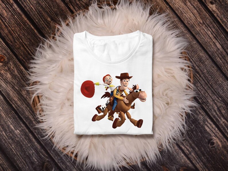 May include: White t-shirt featuring a colorful graphic of Woody and Jessie from Toy Story riding a horse. The design includes a red lasso and the characters in their signature outfits. The shirt is displayed on a fluffy white surface.