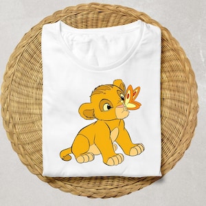 May include: White t-shirt featuring a cartoon lion cub with a butterfly on its nose. The lion cub is yellow and orange, with a friendly expression. The butterfly is orange and yellow. The shirt is folded on a woven surface.