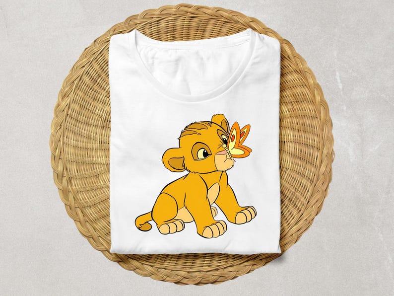 May include: White t-shirt featuring a cartoon lion cub with a butterfly on its nose. The lion cub is yellow and orange, with a friendly expression. The butterfly is orange and yellow. The shirt is folded on a woven surface.