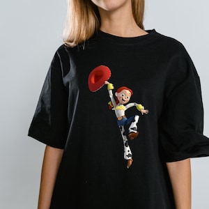 May include: Black t-shirt featuring a colorful graphic of the character Jessie from Toy Story. Jessie is depicted holding a red hat, wearing a white shirt, blue jeans, and cow-print chaps. The shirt has short sleeves.