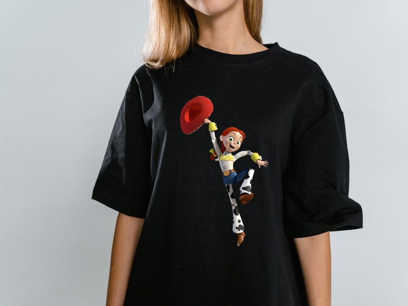 May include: Black t-shirt featuring a colorful graphic of the character Jessie from Toy Story. Jessie is depicted holding a red hat, wearing a white shirt, blue jeans, and cow-print chaps. The shirt has short sleeves.