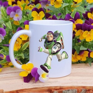May include: White ceramic mug featuring a colorful graphic of Buzz Lightyear from Toy Story in a running pose. The mug is set against a backdrop of yellow and purple pansies, creating a cheerful and playful aesthetic.