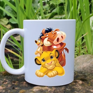 May include: White ceramic mug featuring a colorful illustration of Simba, Timon, and Pumbaa from The Lion King. The mug has a curved handle and is set against a natural background of green foliage and stone.