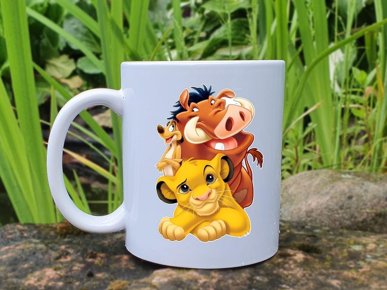 May include: White ceramic mug featuring a colorful illustration of Simba, Timon, and Pumbaa from The Lion King. The mug has a curved handle and is set against a natural background of green foliage and stone.