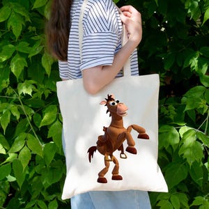 May include: A cream-colored tote bag featuring a cartoon image of a brown horse with a saddle. The horse is in a dynamic pose, with its front legs raised. The bag is held by a person in front of a backdrop of green foliage.