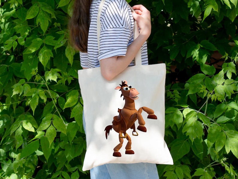 May include: A cream-colored tote bag featuring a cartoon image of a brown horse with a saddle. The horse is in a dynamic pose, with its front legs raised. The bag is held by a person in front of a backdrop of green foliage.