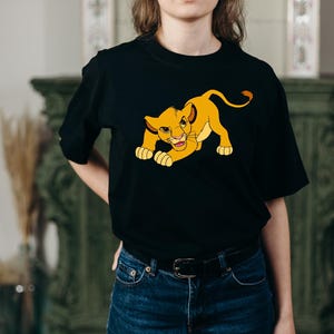 May include: Black t-shirt featuring a cartoon lion cub in yellow and orange. The lion cub is in a crouching position, with its paws on the ground and a playful expression. The shirt is made of a soft material.