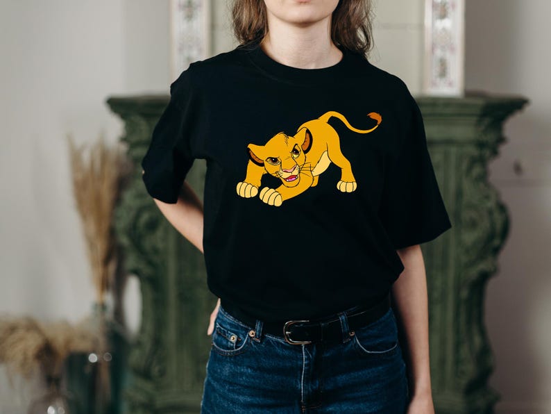 May include: Black t-shirt featuring a cartoon lion cub in yellow and orange. The lion cub is in a crouching position, with its paws on the ground and a playful expression. The shirt is made of a soft material.