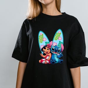 Może przedstawiać: Czarny t-shirt z kolorową grafiką Lilo i Stitch z deskami surfingowymi. Lilo ma na sobie czerwoną i białą sukienkę, a Stitch jest niebieski. Deski surfingowe są jasnoniebieskie z kwiatowymi wzorami.
