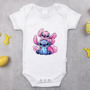 Może przedstawiać: Białe body dla dziecka z kresk&oacute;wkową ilustracją dw&oacute;ch postaci, jednej niebieskiej i jednej r&oacute;żowej, z kwiatkiem. Body ma kr&oacute;tkie rękawy i trzy zatrzaski. W tle dekoracje wielkanocne.