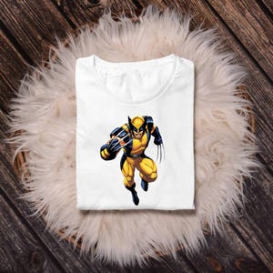 Pu&ograve; includere: T-shirt bianco con grafica a colori di Wolverine nel suo costume giallo e blu. Il supereroe &egrave; raffigurato in una posa dinamica, con gli artigli estesi. La maglietta &egrave; piegata e appoggiata su una superficie bianca e soffice.