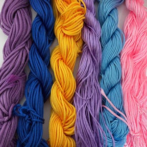 May include: Close-up of colorful nylon cord in shades of purple, blue, yellow, and pink. The cords are tightly wound and appear smooth, ideal for crafting or jewelry making. The vibrant colors create a visually appealing display.