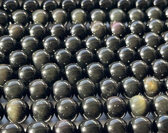 May include: Close-up of a collection of polished, round, black obsidian beads. The beads have a reflective, glossy surface with subtle variations in color, including hints of gold and green. The beads are strung together, suggesting they are for jewelry making.