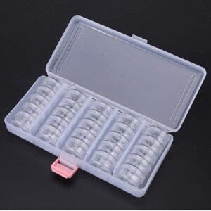 May include: A clear plastic storage box with a hinged lid and a pink clasp. The box is divided into five compartments, each filled with small, clear, round containers. The containers are likely for storing small items like beads or craft supplies.