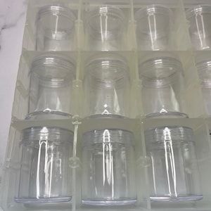 May include: A clear plastic container set, featuring multiple small, cylindrical jars with screw-on lids. The jars are arranged in a grid within a clear plastic tray, designed for organization and storage of small items.