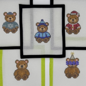 May include: Five white needlepoint canvases featuring brown teddy bears. One bear wears a blue sweater with a Star of David, one a blue hat and coat, one a Santa suit, one a party hat with 2026 glasses, and one is plain. Some have black or green borders.