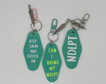 May include: Three teal keychains with white text and metal hardware. The keychains have different phrases: "KEEP CALM AND STITCH ON", "CAN I BRING MY NDLPT", and "NDLPT". Each keychain has a different colored clasp.