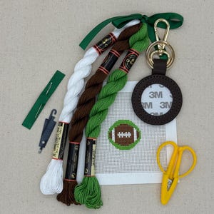 May include: A needlepoint kit featuring a white canvas with a football design, three skeins of embroidery floss in white, brown, and green, a dark brown leather keychain, a green needle threader, a needle, and yellow embroidery scissors.