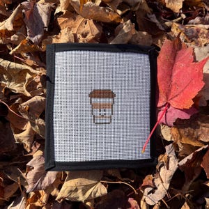 May include: A square, white needlepoint with a black border features a cross-stitched coffee cup design. The cup is brown with white details. The needlepoint is surrounded by fallen autumn leaves.