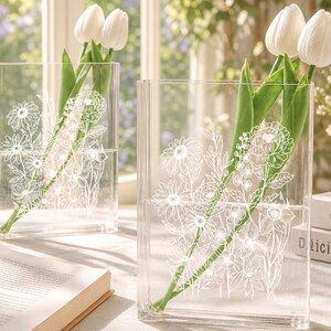 Pode incluir: Dois vasos retangulares de vidro transparente com ilustrações de linhas brancas de várias flores silvestres. Cada vaso contém dois caules de tulipas brancas com folhas verdes. Os vasos estão posicionados sobre uma superfície.