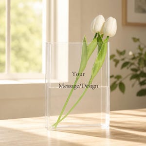 Op de afbeelding: Een heldere rechthoekige vaas met water en drie witte tulpen. De vaas heeft de tekst "Your Message/Design" erop gedrukt. De vaas staat op een houten oppervlak.