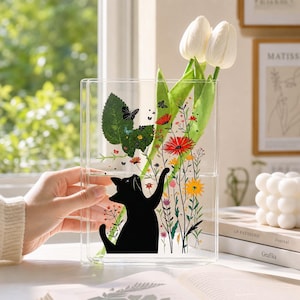 May include: A clear rectangular glass vase featuring a black cat silhouette reaching toward colorful printed wildflowers and butterflies. Two white tulip stems are placed inside the vase, which is positioned on a surface near a window.